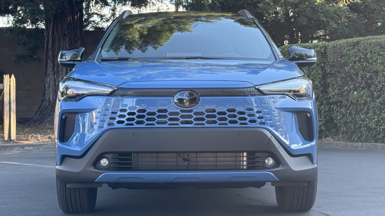 Un plan frontal d'un hybride Corolla Cross Blue montrant sa calandre de couleur corporelle garée devant un buisson